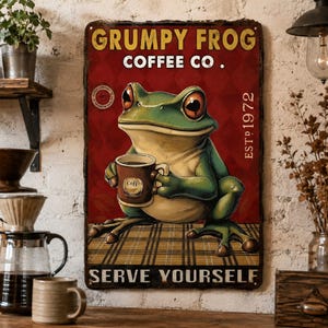 May include: A rectangular metal sign featuring a green frog holding a brown coffee mug. The text reads GRUMPY FROG COFFEE CO. EST 1972 and SERVE YOURSELF. The background is a deep red with a diamond pattern and a plaid surface beneath the frog.