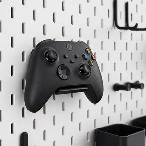 May include: A black wireless video game controller mounted on a white pegboard wall organiser. The controller features two analogue sticks, a directional pad, and four coloured action buttons labelled A, B, X, and Y, positioned below the Xbox logo.