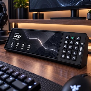 May include: A black Stream Deck control console featuring a wide horizontal digital display showing time, date, and system performance metrics alongside a grid of fifteen square programmable buttons with various white icons for computer control.