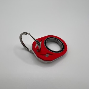 May include: A red and white keyring featuring a circular metal bearing embedded in the centre. The red plastic component has a cutout lightning bolt shape and a silver metal split ring attached through a hole at the top of the keyring.