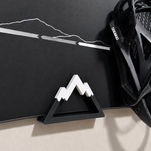 May include: A black snowboard featuring a white mountain range graphic and a white arrow line design. A black Burton snowboard binding is attached to the right side. A small, black and white mountain-shaped plastic object is mounted on the board.