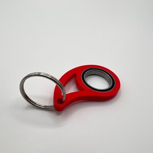 May include: A red teardrop-shaped keyring featuring a central circular metal ball bearing with a black inner ring. A silver-coloured metal split ring is attached to the narrow end of the red frame, providing a functional loop for keys.