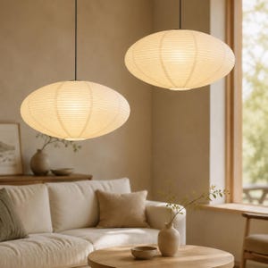 May include: Two off-white, oval-shaped pendant lamps with a ribbed paper texture suspended from the ceiling. The lamps emit a warm, soft glow, featuring a horizontal wire frame structure visible through the thin, translucent material.