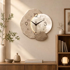 Peut inclure: Une horloge murale avec un cadre beige à texture de pierre irrégulière et un cadran circulaire illuminé rappelant la lune. Elle possède des aiguilles noires pour les heures et les minutes, une trotteuse fine et des chiffres noirs de 1 à 12.