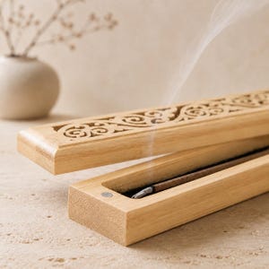 Può includere: Un bruciaincenso in legno color marrone chiaro con coperchio incernierato caratterizzato da intricati motivi a volute tagliati al laser. La scatola è aperta e rivela un bastoncino di incenso acceso che emette un sottile filo di fumo.