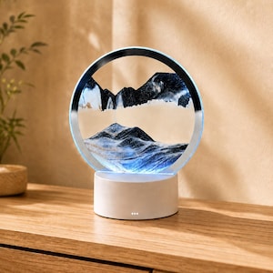 May include: A circular sand art display featuring a white base and a glass frame containing layers of black, white, and blue sand that form mountain-like shapes. The frame is illuminated with a blue light, creating a glowing effect around the sand.