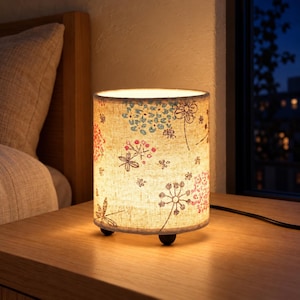 Peut inclure: Une lampe de table cylindrique avec un abat-jour en tissu beige orné d'un motif imprimé de fleurs, de tiges et de libellules bleues, roses et brunes. La lampe est éclairée de l'intérieur et repose sur trois petits pieds arrondis foncés.