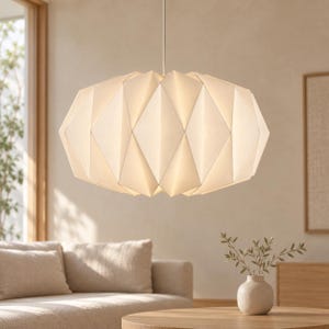 Op de afbeelding: Een witte hanglamp met een geometrische, gefacetteerde, op origami geïnspireerde kap. De lamp is voorzien van een reeks driehoekige panelen die een gestructureerde, veelzijdige vorm creëren, opgehangen aan een dun, recht snoer aan het plafond.