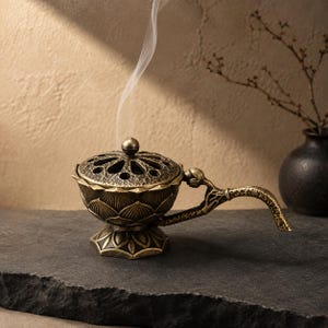 May include: A metallic bronze-toned incense burner shaped like a lotus flower with a perforated lid and a long, textured handle. A thin trail of white smoke rises from the centre of the lid, which features a small spherical knob for lifting.