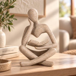 Peut inclure: Une sculpture abstraite beige mouchetée représentant une silhouette assise, la tête appuyée sur une main, tenant un livre ouvert. La silhouette présente une tête et des membres arrondis et lisses, avec une finition texturée rappelant la pierre.