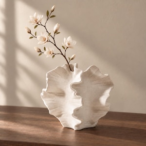 Op de afbeelding: Een witte keramische vaas met een gestructureerd, golvend, schelpachtig ontwerp met een enkele tak met verschillende witte magnolia bloemen en groene bladeren. De vaas heeft een gespikkelde afwerking en een brede, golvende rand met plooien.