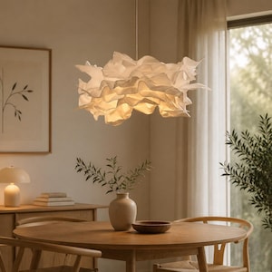 May include: A white pendant light fixture featuring a layered, ruffled, cloud-like shade suspended from a thin cord. The light emits a warm, soft glow from within the intricate, undulating folds of the material, creating a textured appearance.