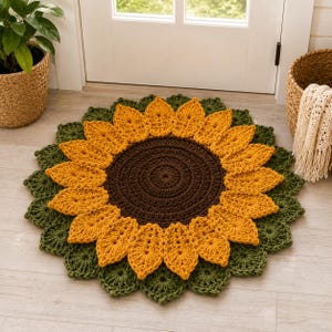 May include: A round, crocheted rug shaped like a sunflower. The center is a dark brown circle, surrounded by two layers of bright yellow, petal-shaped stitches. The outer edge features a border of dark green, leaf-shaped crochet motifs.
