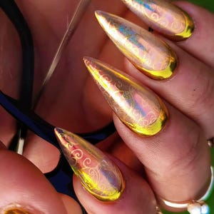 May include: Stiletto-shaped artificial nails featuring a reflective iridescent finish with gold-toned filigree patterns and solid gold-coloured tips. The nails are long and pointed, displaying a metallic sheen that catches the light.