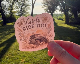 May include: A die-cut sticker featuring a light peach background with a dark brown illustration of an off-road vehicle, mountains, a checkered flag, flowers, and a butterfly. The text Girls RIDE TOO is printed in a dark brown script and serif font.