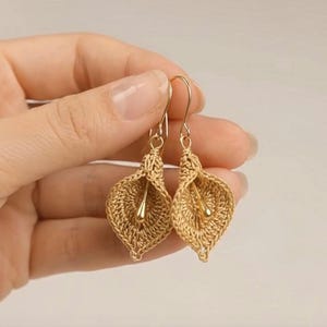 Puede incluir: Un par de pendientes de ganchillo en tono dorado con forma de lirios cala. Cada pendiente presenta un diseño de pétalo tejido y texturizado con un estambre liso en forma de gota de oro metálico suspendido en el centro, unido a un gancho de alambre dorado.