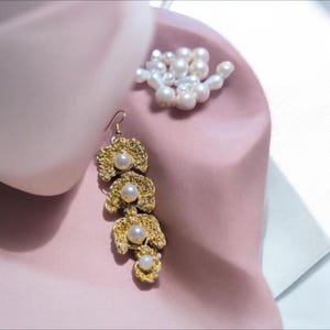 Peut inclure: Une longue boucle d'oreille crochetée de couleur dorée ornée de quatre perles blanches sphériques disposées en ligne verticale. La boucle d'oreille possède une attache à crochet. Un petit groupe de perles blanches est visible à proximité.