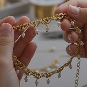May include: A gold-colored crocheted choker necklace featuring a delicate openwork pattern and small white spherical beads suspended from the bottom edge. The necklace includes a metal chain extender with circular links at the end for adjustable sizing.
