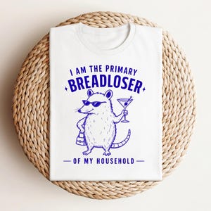 May include: A white t-shirt featuring a blue graphic of an opossum wearing sunglasses, holding a martini glass and a small handbag. The text reads I AM THE PRIMARY BREADLOSER OF MY HOUSEHOLD in blue ink, centred on the front of the garment.