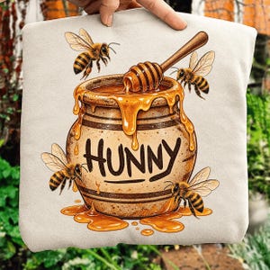 May include: A beige sweatshirt featuring a graphic illustration of a tan ceramic honey pot with the word HUNNY printed in black. A wooden honey dipper rests inside the pot, with golden honey dripping down the sides, surrounded by four bees.
