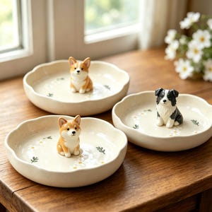 May include: Three round, cream-colored ceramic trinket dishes with scalloped edges. Each dish features a small, hand-painted dog figurine in the center: two are brown and white corgis, and one is a black and white border collie. White daisy motifs.