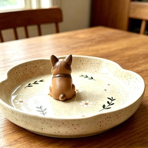 May include: A round, cream-colored ceramic trinket dish with a speckled finish, featuring a small, brown, sculpted dog figure sitting in the center. The dish is decorated with painted white daisy flowers and green leafy sprigs on the interior surface.