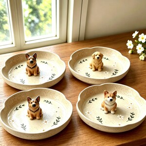 May include: Four round, speckled cream-colored ceramic dishes, each featuring a small, hand-painted dog figurine at the center. The dishes are decorated with delicate green leaf sprigs and small white flowers painted on the interior base.