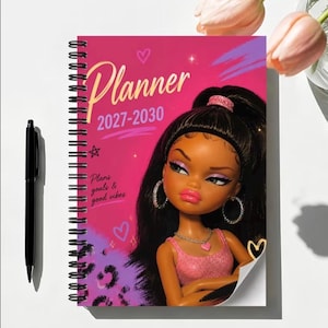 May include: A spiral-bound planner with a pink cover featuring the text Planner 2027-2030 and Plans goals & good vibes. The cover displays an illustration of a doll with long black hair, wearing a pink top, hoop earrings, and a heart necklace.