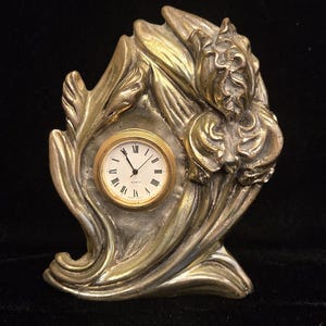May include: A gold-toned metal table clock featuring a sculpted floral design with large petals and leaves surrounding a circular clock face. The clock face has a white background, black Roman numerals, and black hands with the word Quartz printed.