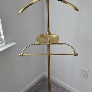 May include: A polished brass valet stand featuring a coat hanger top with a spherical finial, a central oval tray for small items, and a lower horizontal bar for trousers. The structure is supported by a circular base with a decorative rope edge.