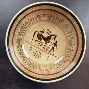 May include: A round ceramic bowl with a beige speckled base and dark brown rim. The interior features a dark brown graphic illustration of a horse, a bird, and a human figure, surrounded by concentric rings and small geometric patterns in brown and red.
