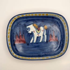 May include: A rectangular ceramic tray with rounded corners, featuring a deep blue background, a central white horse figure with brown and red accents, two red triangular shapes, and a yellow dotted border along the rim of the dish.