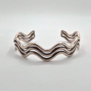 May include: A silver-toned metal cuff bracelet featuring a triple-layered wave design. The piece consists of three parallel, undulating wire strands that are connected at the ends, creating a continuous, flowing, and rhythmic pattern throughout.