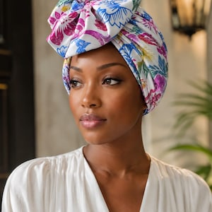 May include: A head wrap featuring a white base with a vibrant floral pattern in shades of blue, pink, and green. The fabric is gathered at the top into a large, structured knot detail, providing full coverage around the head with a smooth finish.