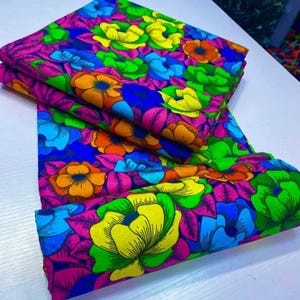 May include: A folded piece of fabric featuring a vibrant floral pattern with large, stylized flowers in bright shades of yellow, orange, blue, and green against a deep magenta background, with dark outlines defining each petal and leaf shape.