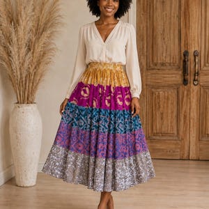 May include: A long-sleeved cream-coloured blouse with a V-neckline paired with a high-waisted, tiered midi skirt. The skirt features horizontal bands of mustard yellow, magenta, teal, purple, and grey, each decorated with intricate, repeating patterns.