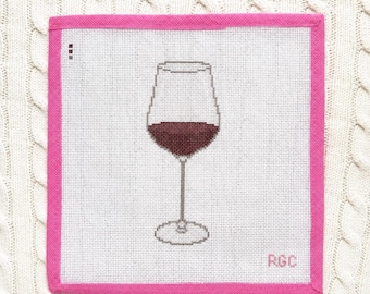 Peut inclure: Une toile de canevas carrée représentant un verre à vin centré rempli de vin rouge foncé. La toile présente une bordure en tissu rose vif et les lettres RGC brodées dans le coin inférieur droit sur un fond en maille quadrillée blanche.