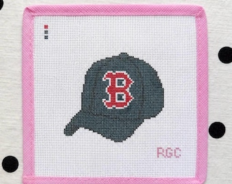 Peut inclure: Une toile de canevas carrée représentant une casquette de baseball gris foncé avec une lettre B rouge au centre. La toile présente une bordure en tissu rose clair et les lettres RGC brodées en bas à droite, avec un petit guide de points rouge et gris.