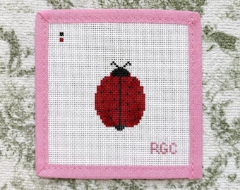 May include: A square needlepoint canvas featuring a red ladybug with black spots and a black head centered on a white grid. The canvas is framed by a pink fabric border and includes the letters RGC in the bottom right corner and two small color squares in the top left corner.