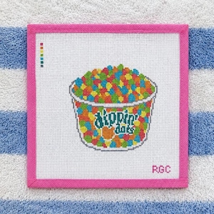 May include: A square needlepoint canvas featuring a colorful cross-stitch design of a cup filled with multi-colored Dippin' Dots. The design includes the text 'dippin' dots' in teal, a small color key in the top left, and the initials 'RGC' in the bottom right, all framed by a bright pink fabric border.