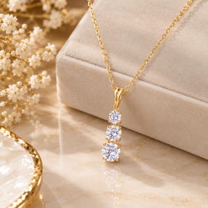 May include: A gold necklace featuring a vertical pendant with three round, clear, faceted stones arranged in a descending size order. The pendant is suspended from a delicate gold chain, showcasing a bright, reflective metallic finish.