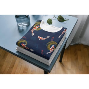 Peut inclure: Un t-shirt bleu marine plié présentant un motif répétitif de poissons koï orange, blancs et noirs nageant parmi des motifs de vagues circulaires dorés. Le vêtement possède un col rond blanc uni et est empilé sur d'autres t-shirts pliés.