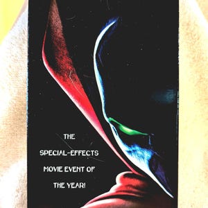 Könnte beinhalten: Eine rechteckige VHS-Kassette für den Film Spawn. Das schwarze Cover zeigt den Titel SPAWN in blauen Buchstaben oben, ein stilisiertes Charakterprofil mit einem leuchtend grünen Auge und den Text THE SPECIAL-EFFECTS MOVIE EVENT OF THE YEAR!