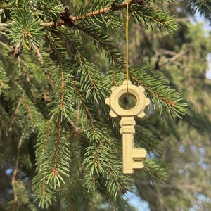 May include: A gold-coloured key ornament with a gear-shaped bow and a textured surface, suspended by a thin gold string. The key features a cylindrical shaft and a stepped bit, hanging from a green pine branch with visible needles and cones.