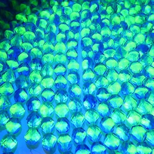 Può includere: Una collezione di numerose piccole perle di vetro traslucide e sfaccettate disposte a griglia. Le perle mostrano una lucentezza vibrante con tonalità alternate di verde neon brillante e blu elettrico intenso, creando un effetto luminoso.