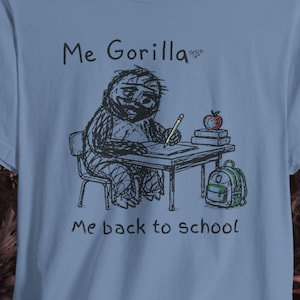 May include: A light blue t-shirt featuring a black line drawing of a gorilla sitting at a desk writing with a pencil. The graphic includes a red apple, a stack of books, a green backpack, and the text "Me Gorilla" and "Me back to school".