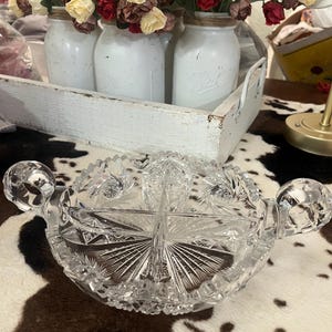 May include: A clear glass divided serving dish featuring a complex starburst and geometric cut pattern. The round dish includes two curved side handles and is partitioned into four distinct sections with a serrated, scalloped outer rim edge.