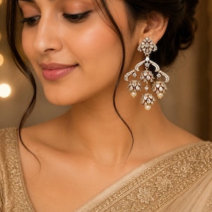 Peut inclure: Une grande boucle d'oreille ornée de style lustre, dotée d'un clou triangulaire incrusté de pierres claires, relié à une structure à plusieurs niveaux composée d'éléments en forme de cloche ornés de pierres blanches et de petites perles crème.