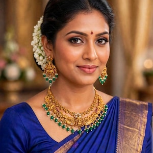 May include: A gold necklace and matching drop earrings featuring intricate metalwork, small red accents, and dangling green beads. The jewelry set is worn with a deep blue garment that has a gold border and white floral hair accessories.