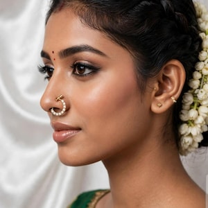 May include: A gold nose ring featuring a circular band adorned with small, clear, sparkling stones. The jewelry is worn in the nostril, showcasing a polished metallic finish and a delicate, symmetrical arrangement of stones along the hoop's curve.